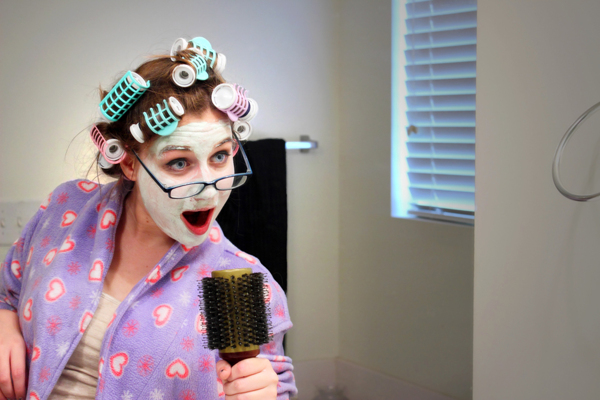 A caucasian girl wearing a colorful robe, curlers, facial mask and glasses sings into a hairbrush in front of the bathroom mirror.